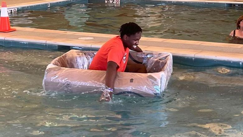 Student paddling cardboard boat in a pool.